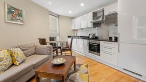 Una cocina moderna con armarios blancos, horno empotrado y microondas. Hay una zona de comedor con una mesa y sillas de madera, y un sofá color beige con cojines estampados.