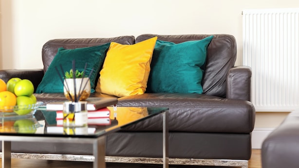 Un sofá de cuero marrón con cojines verde azulado y amarillo, una mesa de centro de vidrio con libros y una planta, y un radiador al fondo.