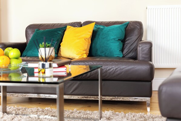 A brown leather sofa with teal and yellow cushions, a glass coffee table with books and a plant, and a radiator in the background.