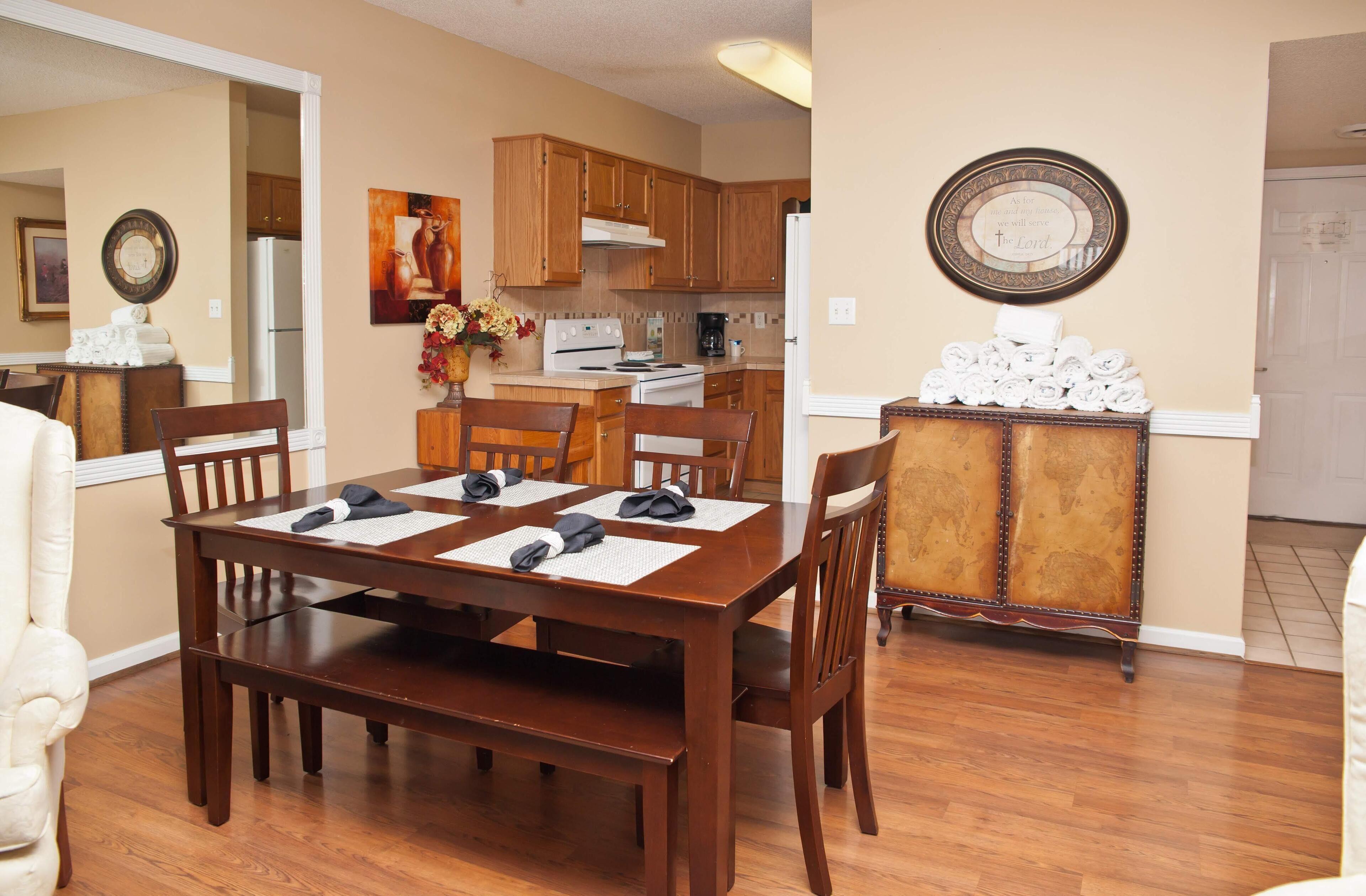 A dining area with a wooden table and chairs, a kitchen in the background, and a decorative plate on the wall.