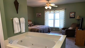 A bedroom with a large bathtub, a bed with a brown blanket, a chair, a television, and a ceiling fan. - Whispering Pines 252 (Pigeon Forge)