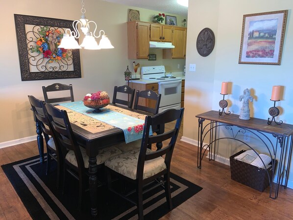A dining area with a wooden table, chairs, a chandelier, and a kitchen in the background.