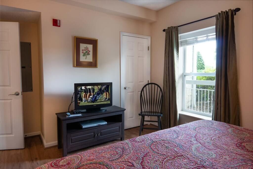 A bedroom with a bed, a television on a stand, a chair, a door, and a window with curtains.