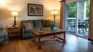Un salon avec un canapé vert, une table basse en bois, deux lampes, un tableau au mur et une porte-fenêtre coulissante donnant sur une terrasse extérieure avec des chaises.