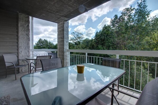A glass table on a balcony with a metal railing, chairs, and a view of trees and sky.
