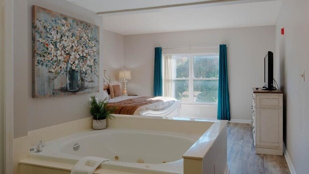 A modern bathroom with a large bathtub, a sink, and a mirror. There is a window with curtains and a bed in the background. - Whispering Pines 333 (Pigeon Forge)