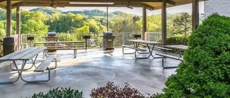 An outdoor covered patio with picnic tables and barbecue grills, surrounded by greenery and a view of hills.