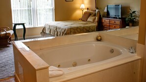 A bedroom with a large bathtub, a bed with patterned bedding, a dresser, a television, and a window with curtains. - Whispering Pines 324 (Pigeon Forge)
