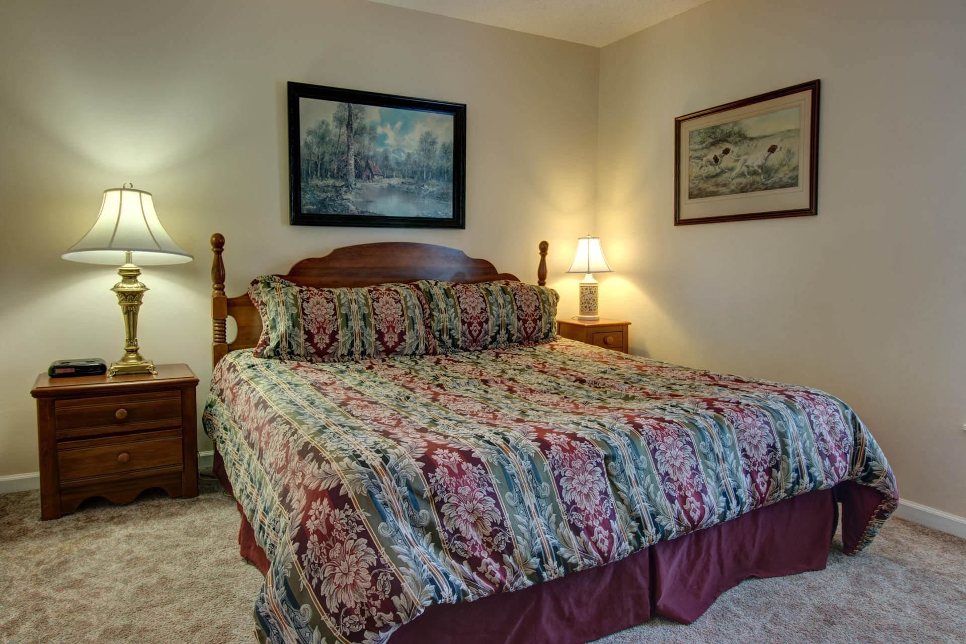A bedroom with a large bed, two bedside tables with lamps, a wooden headboard, and two framed pictures on the walls.