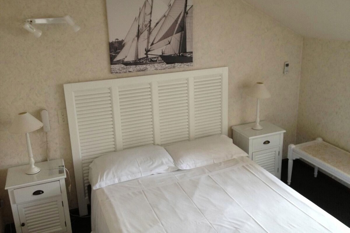 A bedroom with a bed, two bedside tables, a nightstand, a wall-mounted lamp, and a painting of sailboats.