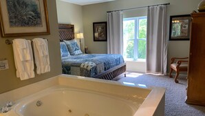 A bedroom with a large bathtub, a bed with patterned bedding, a window with curtains, and a wooden chair. - Whispering Pines 343 (Pigeon Forge)