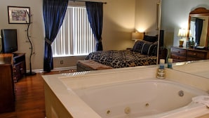 A bedroom with a large bathtub, a bed with a patterned bedspread, a dresser, a television, and a window with curtains. - Whispering Pines 322 (Pigeon Forge)