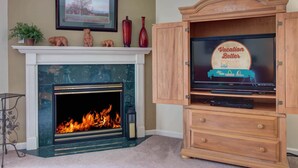 Una chimenea de diseño moderno, un cuadro con un paisaje enmarcado encima y un mueble de madera con un televisor mostrando una imagen temática de vacaciones.