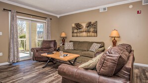 A living room with a brown leather sofa, a matching armchair, a wooden coffee table, and a beige couch. There are two lamps on either side of the sofa, and a painting of trees on the wall. - Whispering Pines 413hc (Pigeon Forge)