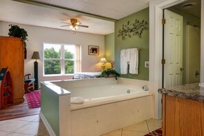 Een badkamer met een groot bad, een plafondventilator, een raam met uitzicht op groen en een deur naar een andere kamer.