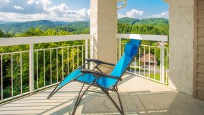 A balcony with a blue lounge chair, white railing, and a view of green mountains. - Whispering Pines 453 (Pigeon Forge)