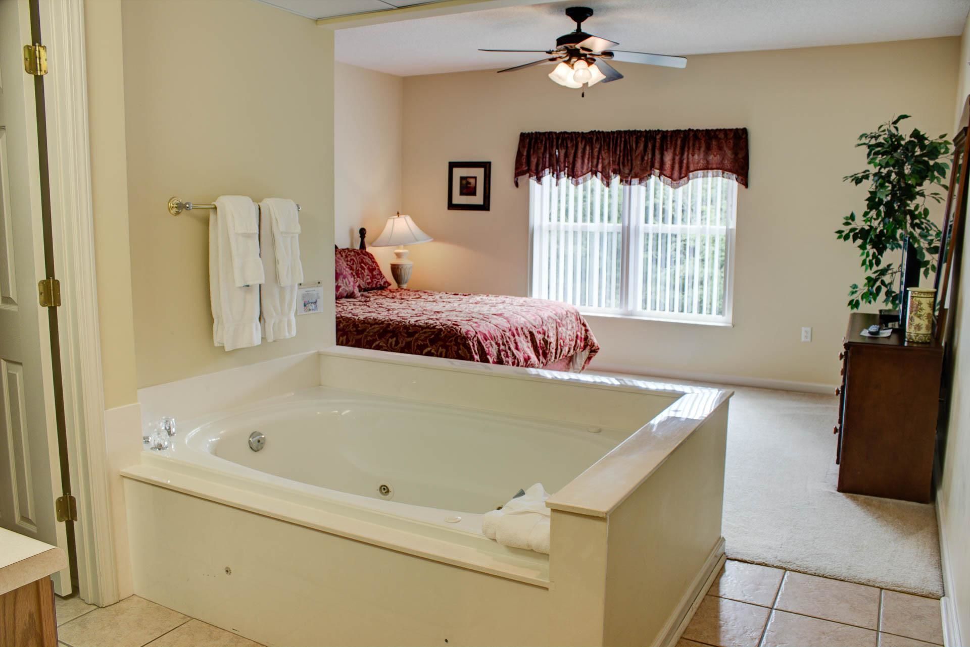 A bathroom with a large bathtub, a bed with a red bedspread, a window with curtains, and a ceiling fan.
