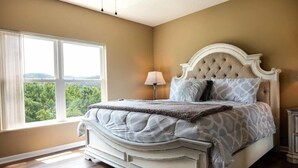 Une chambre avec une grande fenêtre, un lit doté d’une tête de lit capitonnée et une table de chevet avec une lampe.