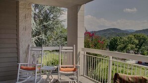 Ein Balkon mit weißen Schaukelstühlen und einem Tisch mit Blick auf eine gebirgige Landschaft mit Bäumen und Blumen.