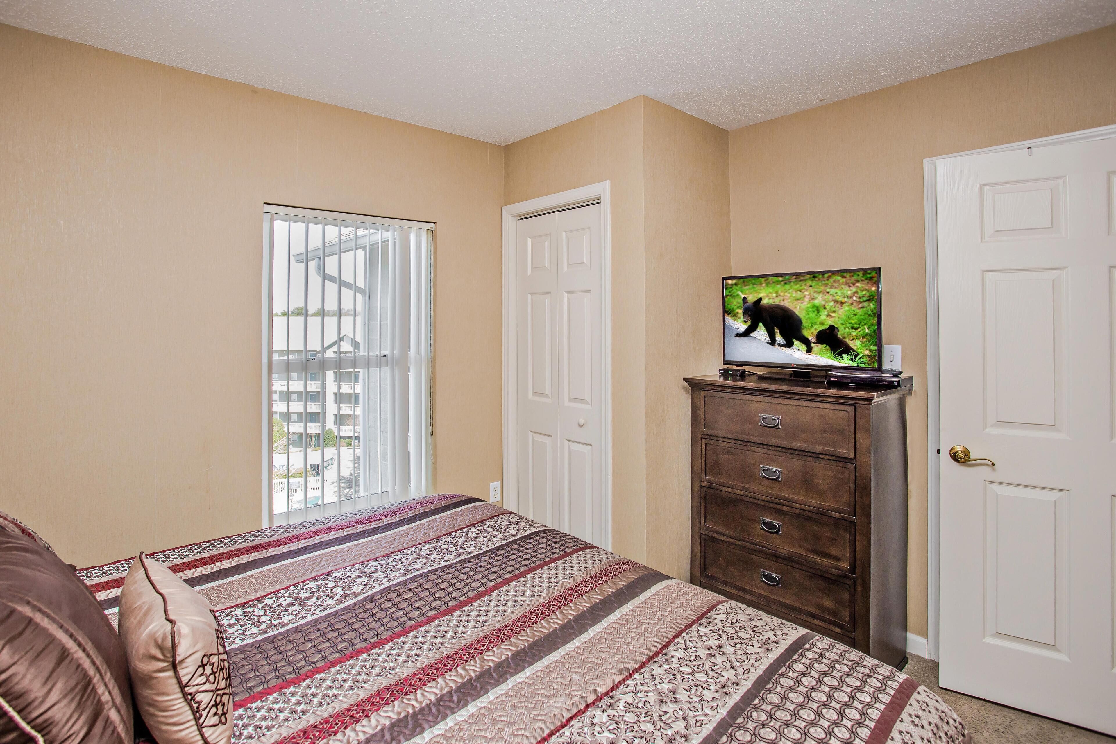 A bedroom with a bed, a dresser, a television, and a window with blinds.