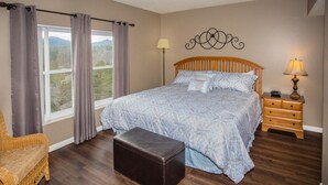 A bedroom with a large bed, a wooden nightstand, a wicker chair, and a window with curtains. - Whispering Pines 541 (Pigeon Forge)