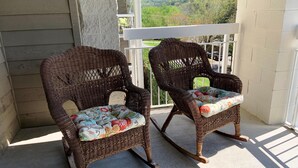 Twee rieten schommelstoelen met bloemenkussens op een balkon met uitzicht op glooiende groene heuvels.