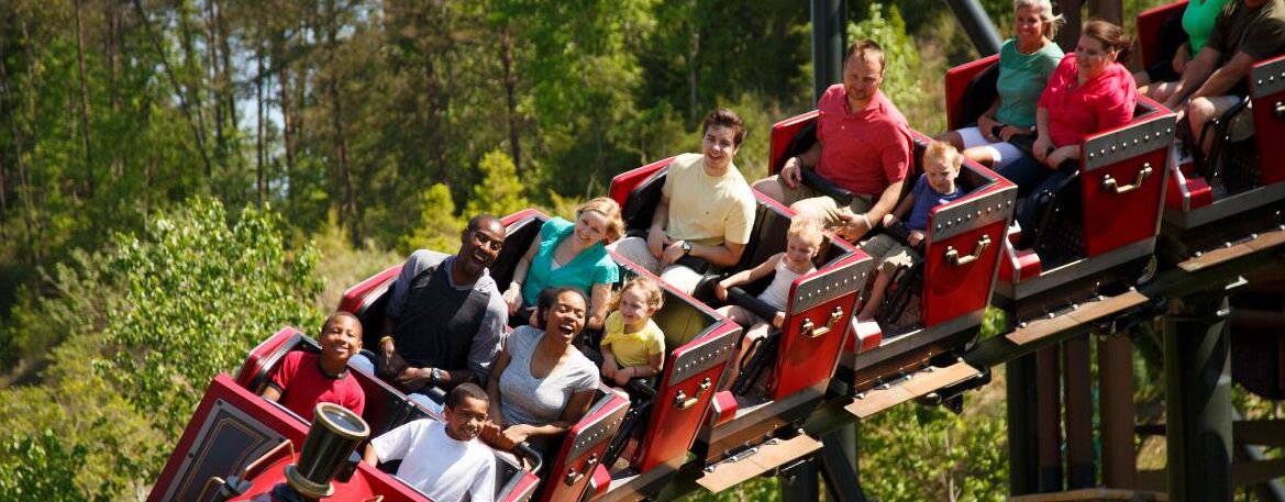 A group of people on a wooden roller coaster with open sides, surrounded by greenery.