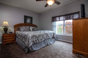 Quarto com cama, mesa de cabeceira, abajur, ventoinha no teto, janela com cortinas e um armário de madeira.