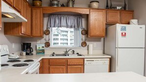 A compact kitchen with wooden cabinets, a white refrigerator, a coffee maker, a dishwasher, and a window with a view of a building. - Whispering Pines 634 (Pigeon Forge)