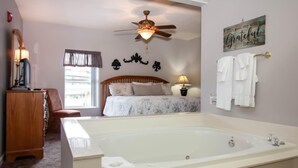 A bedroom with a large bathtub, a ceiling fan, a bed with a wooden headboard, and a window with curtains. - Whispering Pines 634 (Pigeon Forge)