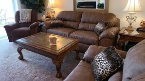 A living room with a brown leather sofa, a glass coffee table, and leopard print cushions. There is a fireplace with a framed mirror above it, and two lamps on side tables. - Whispering Pines 643 (Pigeon Forge)