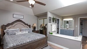 Een slaapkamer met een groot bed, een plafondventilator, een kaptafel met spiegel en een klein raam.
