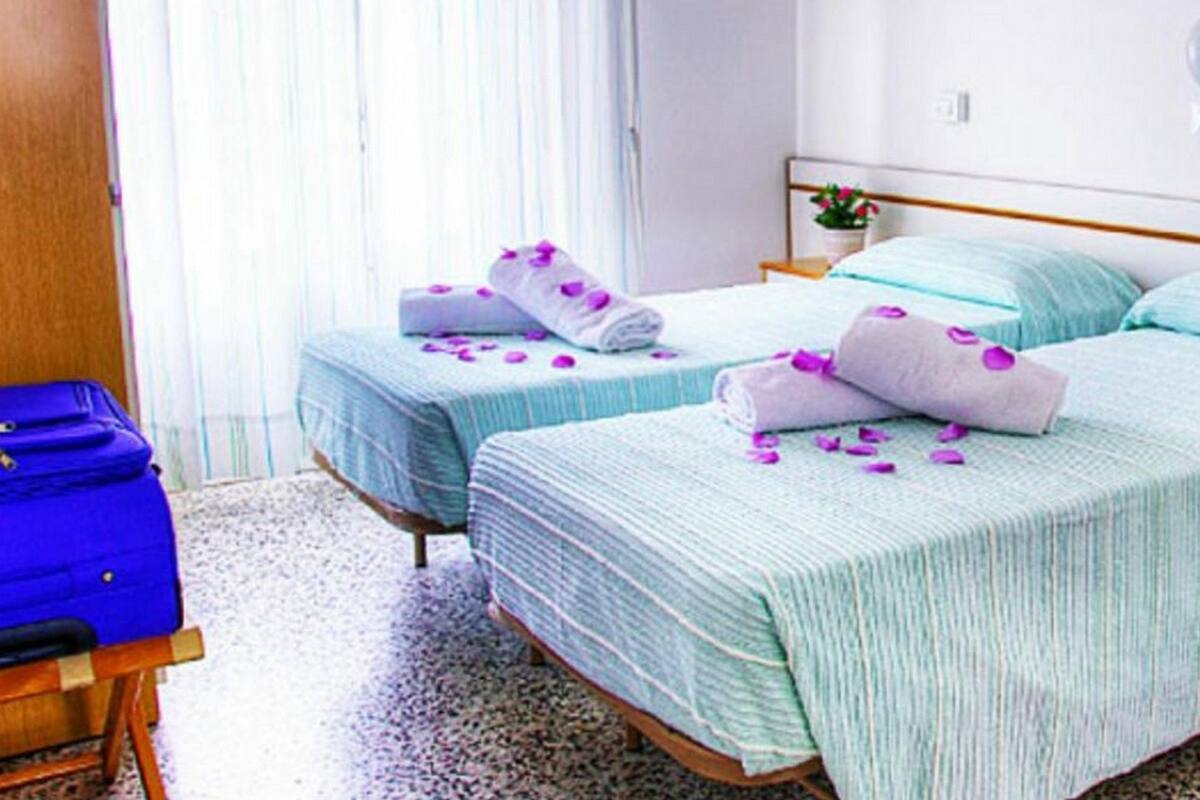 A hotel room with two beds, each with a blue and white striped bedspread, and a blue suitcase on a wooden chair.