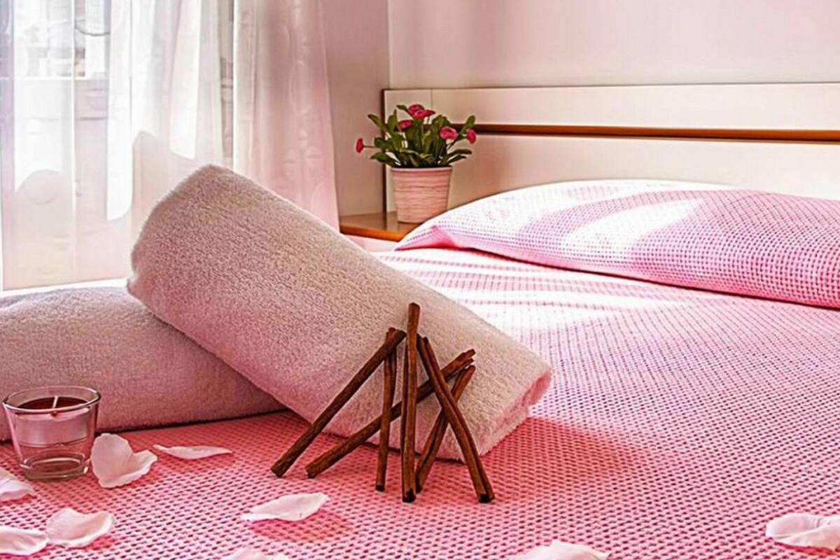 A neatly made bed with pink bedding, a rolled towel, a small glass of water, and a candle on a bedside table.