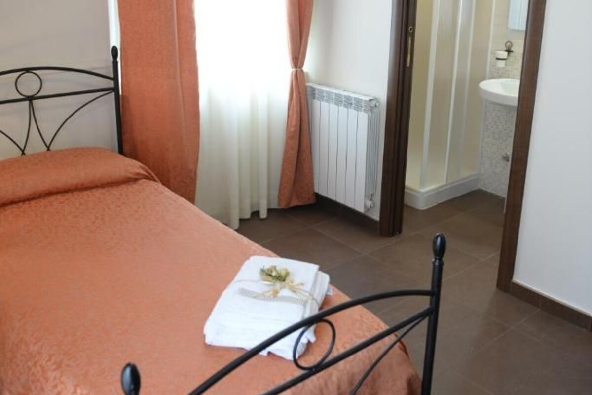 A bedroom with a bed, a window with curtains, a radiator, a bathroom visible through an open door, and a mirror on the wall.