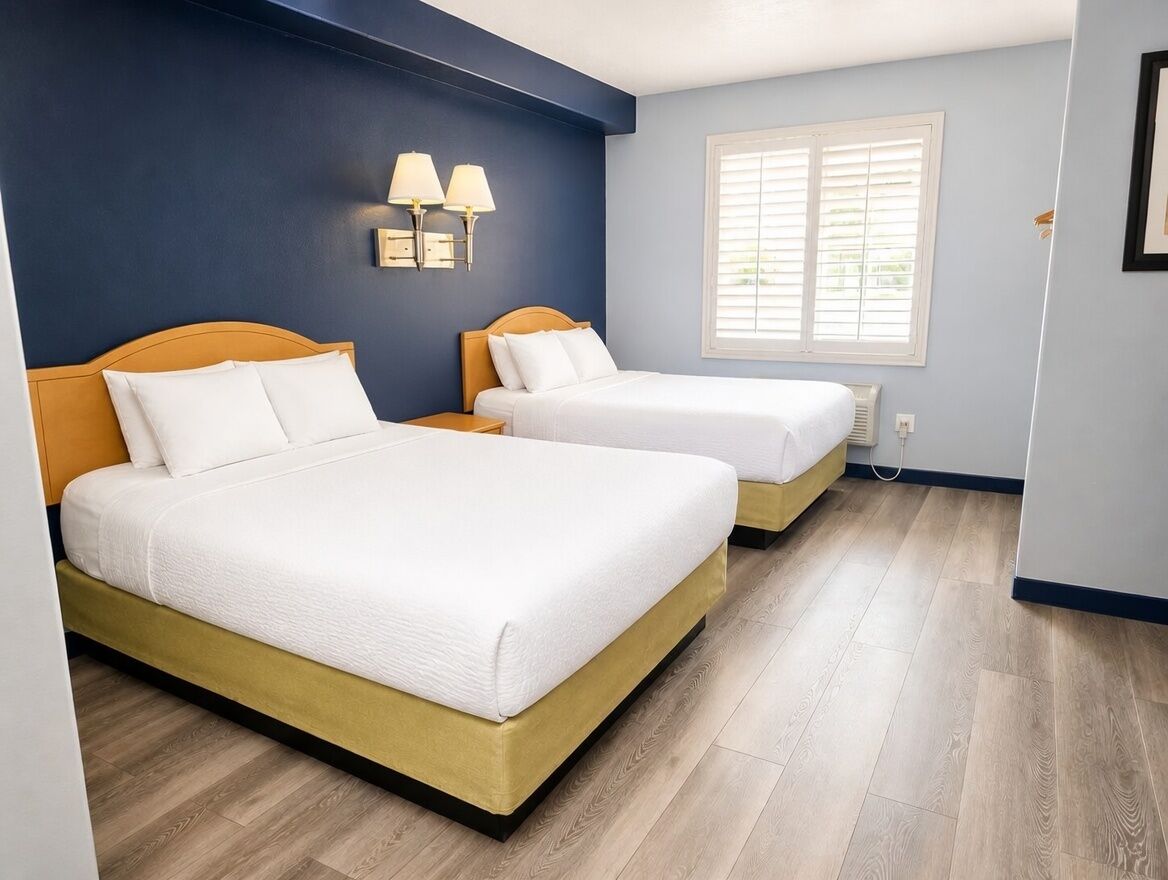 A hotel room with two beds, a window with blinds, a wall-mounted lamp, and a radiator.