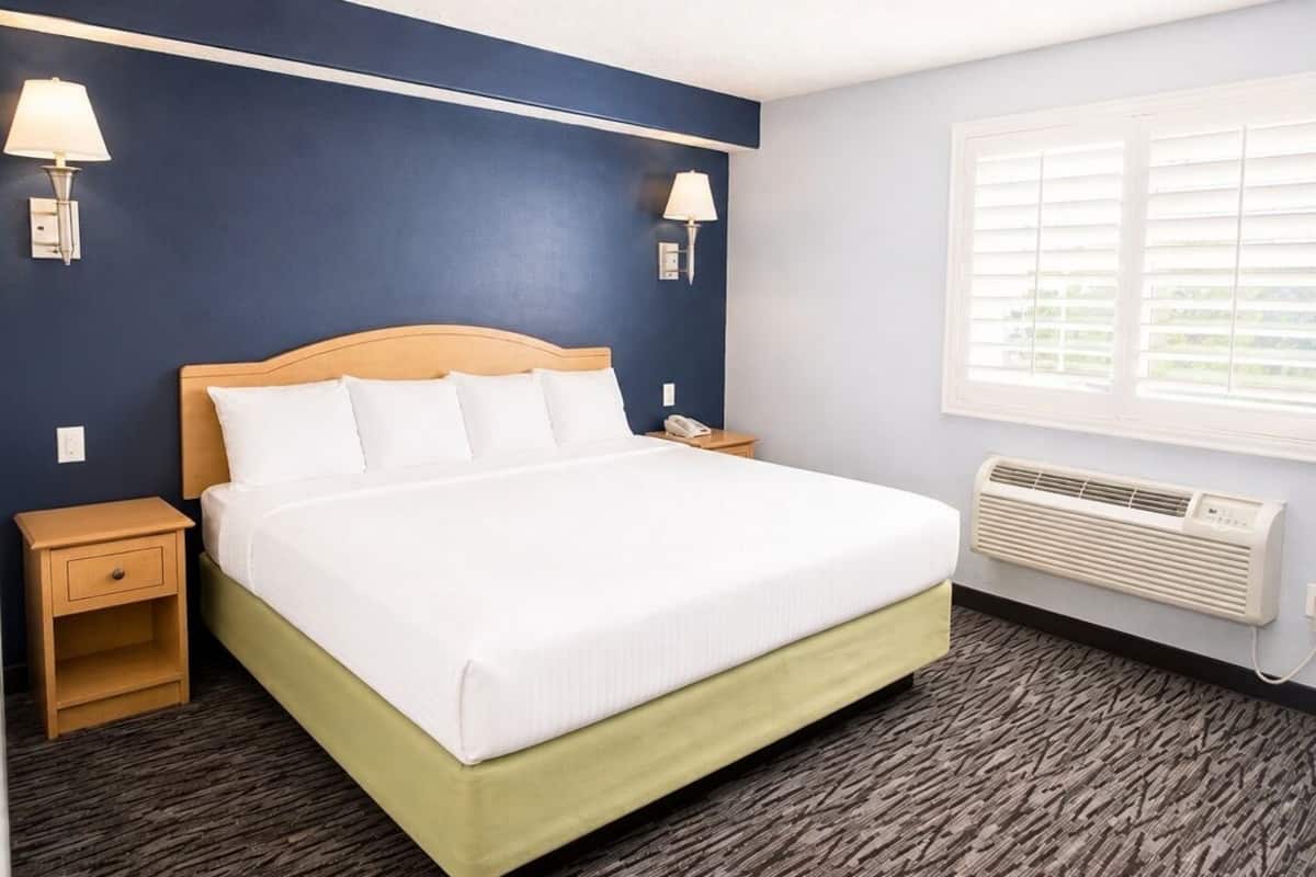 A hotel room with a bed, two bedside lamps, a nightstand, a window with blinds, and a wall-mounted air conditioner.