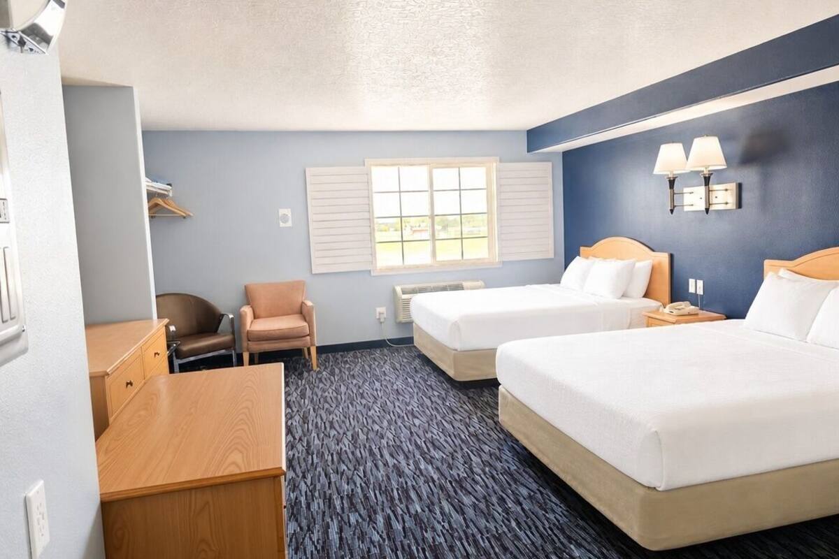 A hotel room with two beds, a desk, a chair, a window, and a wall-mounted lamp.