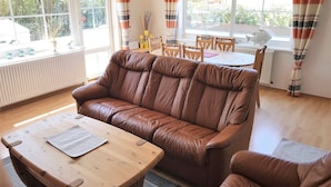 A cozy living room with a brown leather sofa, a wooden coffee table, and a dining area with a table and chairs. - Haus Deichkrone (Dagebuell)