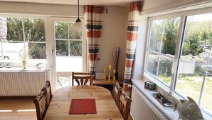 A dining area with a wooden table and chairs, a red placemat, and a view of the garden through large windows. - Haus Deichkrone (Dagebuell)