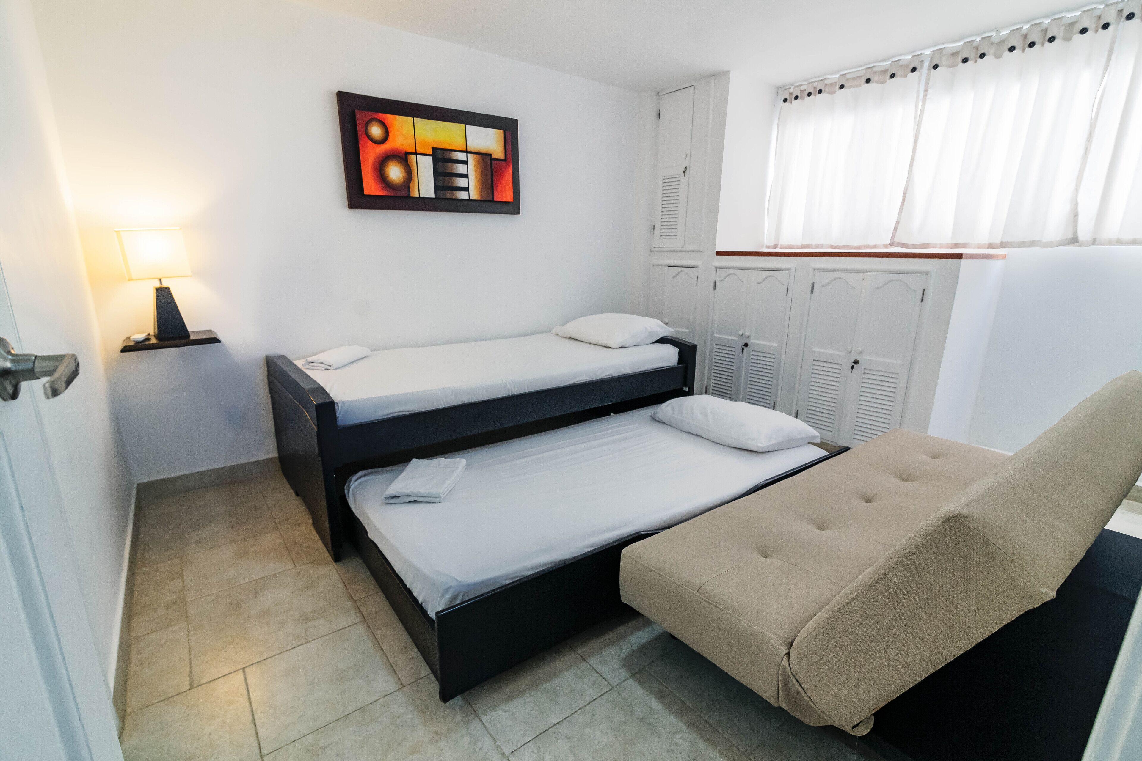 Quarto com duas camas, um sofá, um abajur, um quadro e uma janela com cortinas.