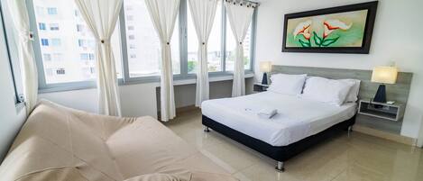 A hotel room with a bed, a sofa, a painting, and a view of buildings outside.