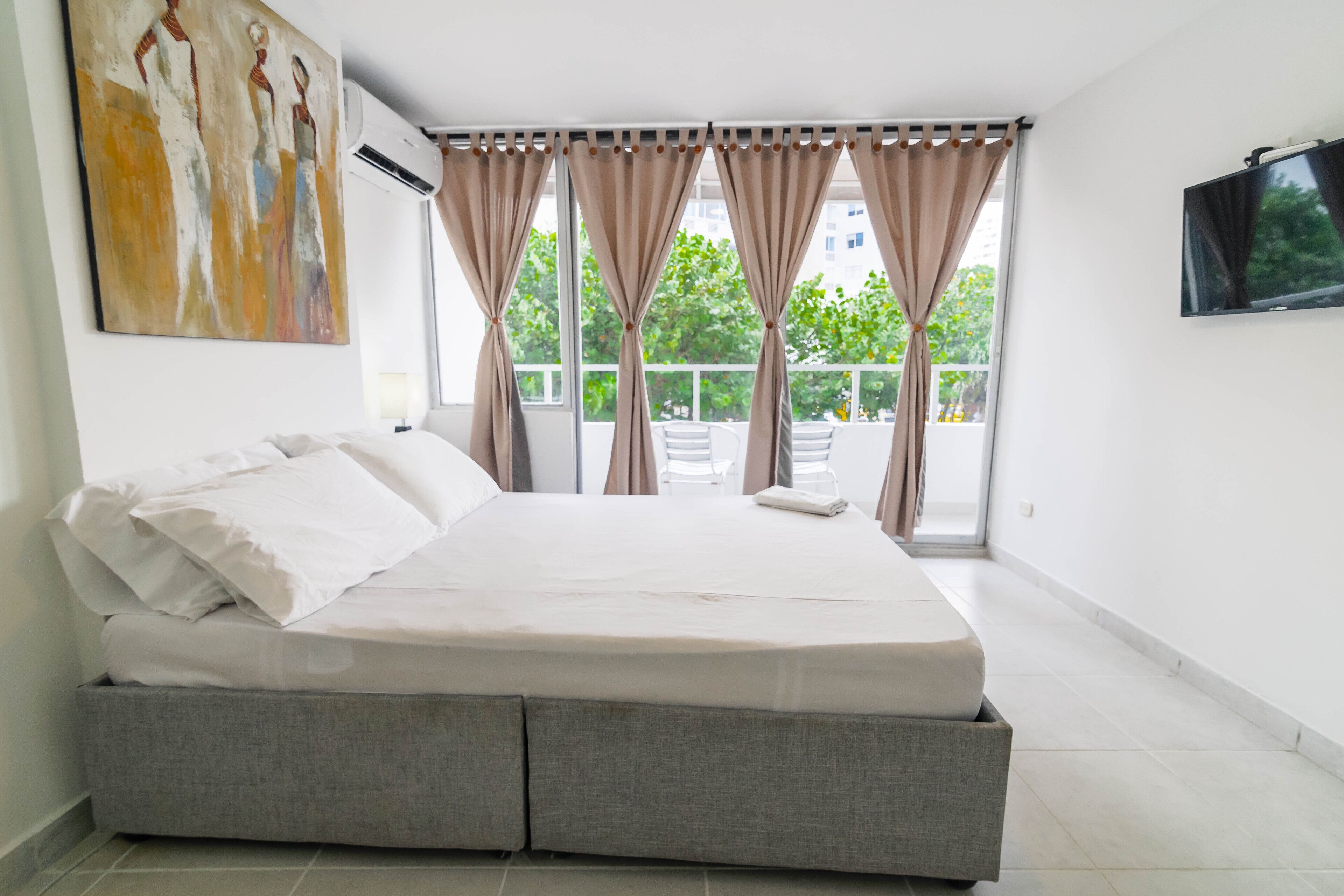 A modern hotel room with a large bed, a flat-screen TV, and a balcony with a view of trees.