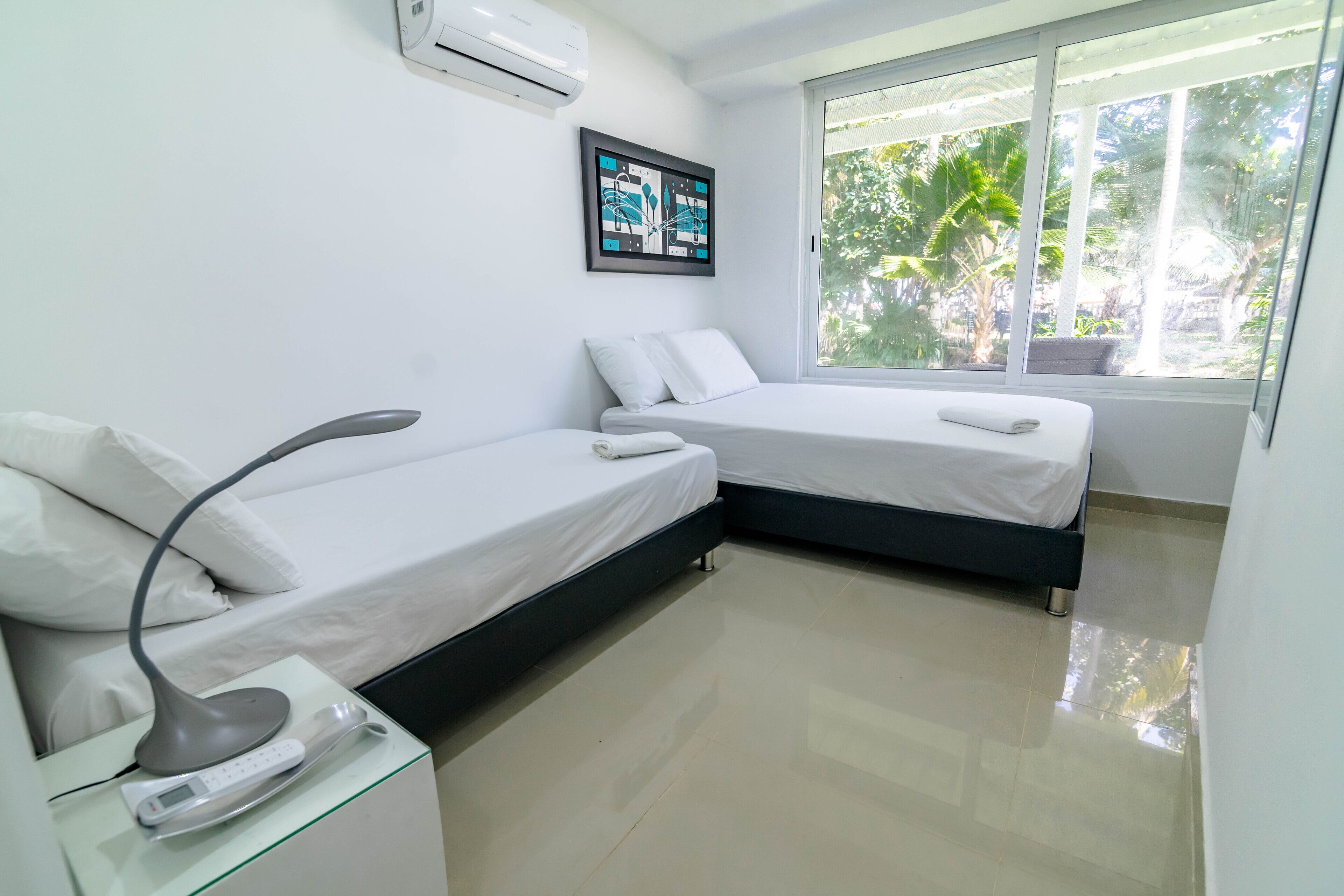 A modern bedroom with two beds, a bedside table with a lamp, a framed picture on the wall, and a window with a view of greenery.