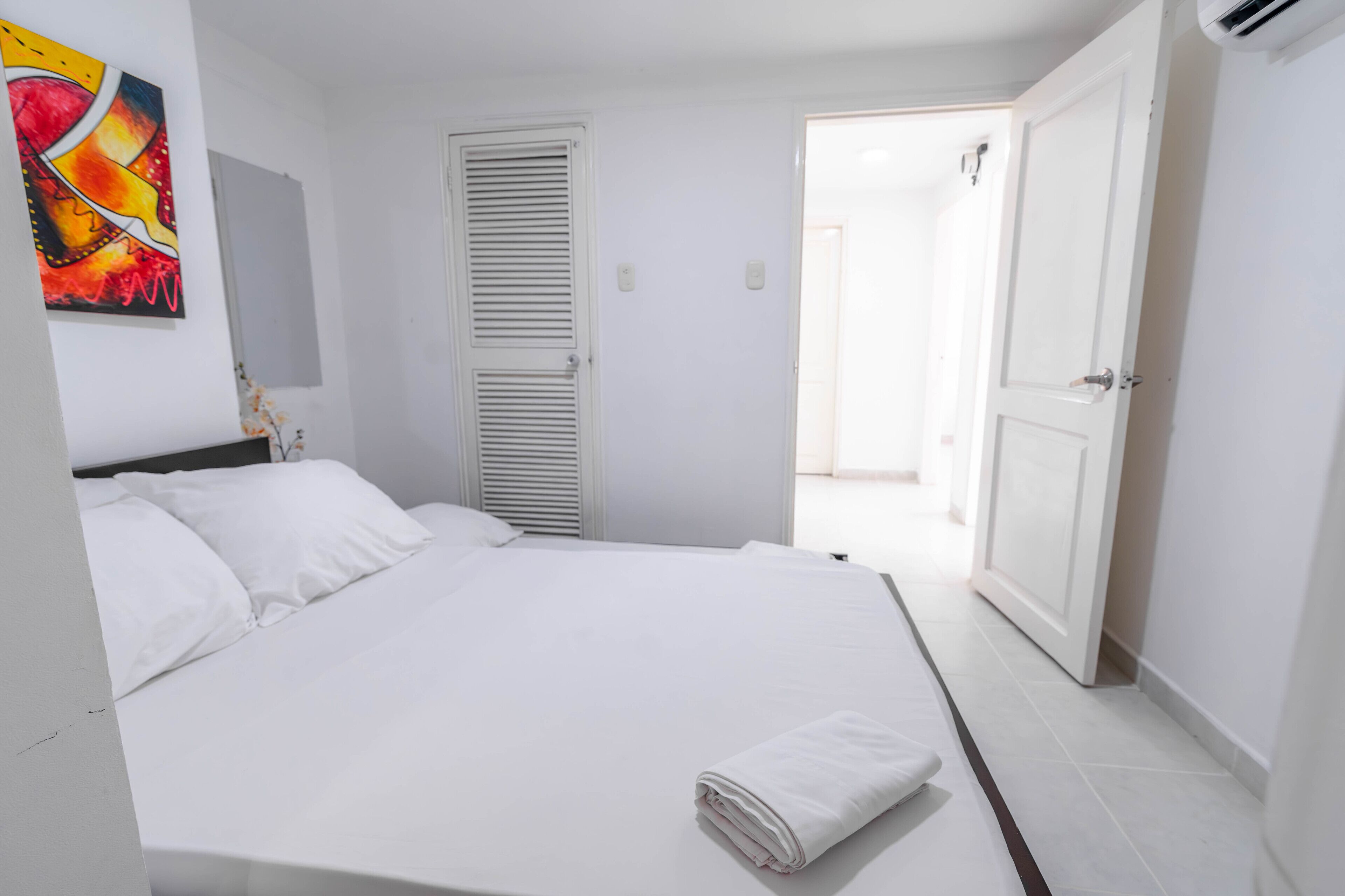 A clean, white room with a bed, a door, and a wall-mounted artwork.