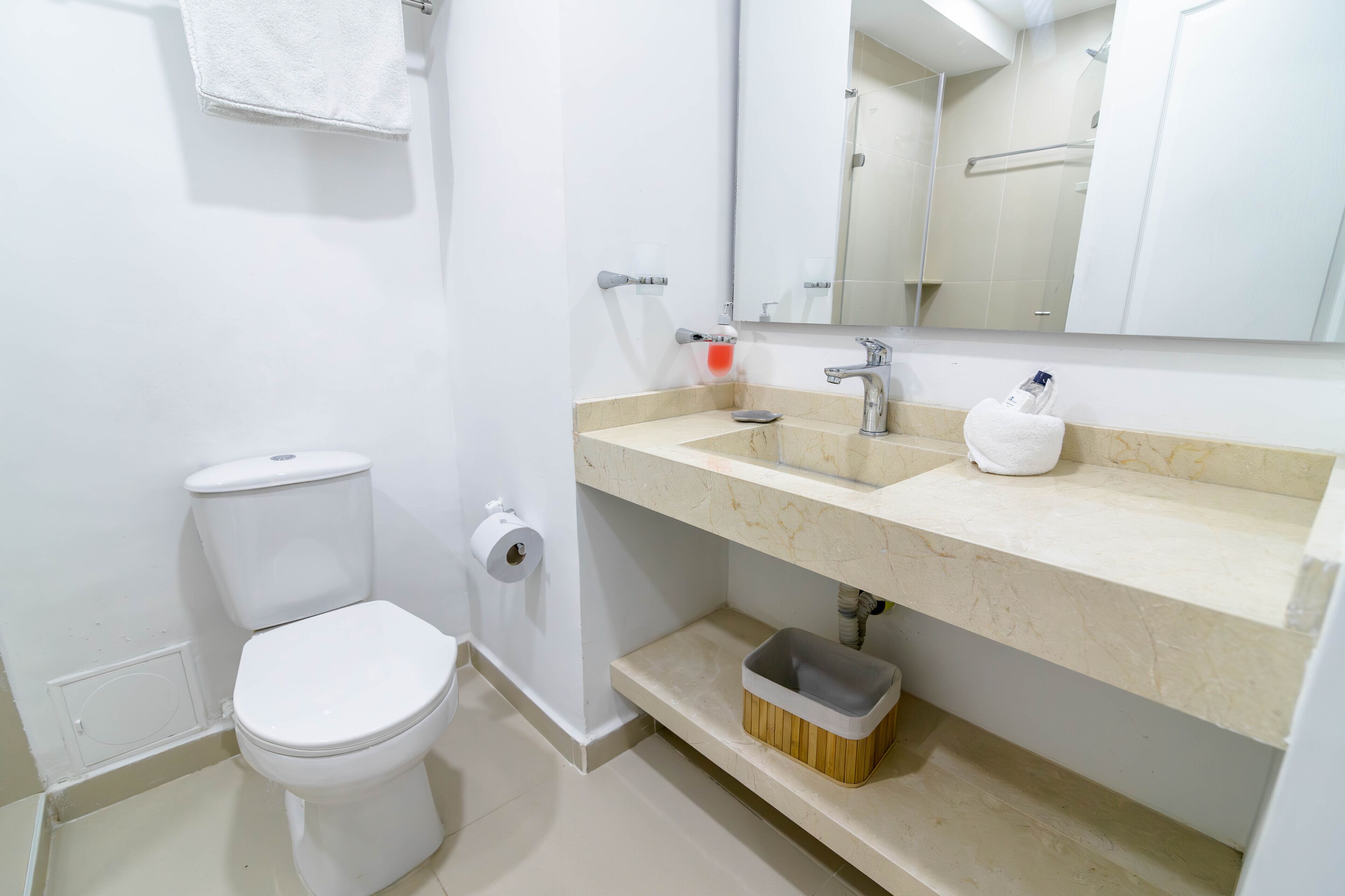 A bathroom with a toilet, sink, mirror, and storage shelf.