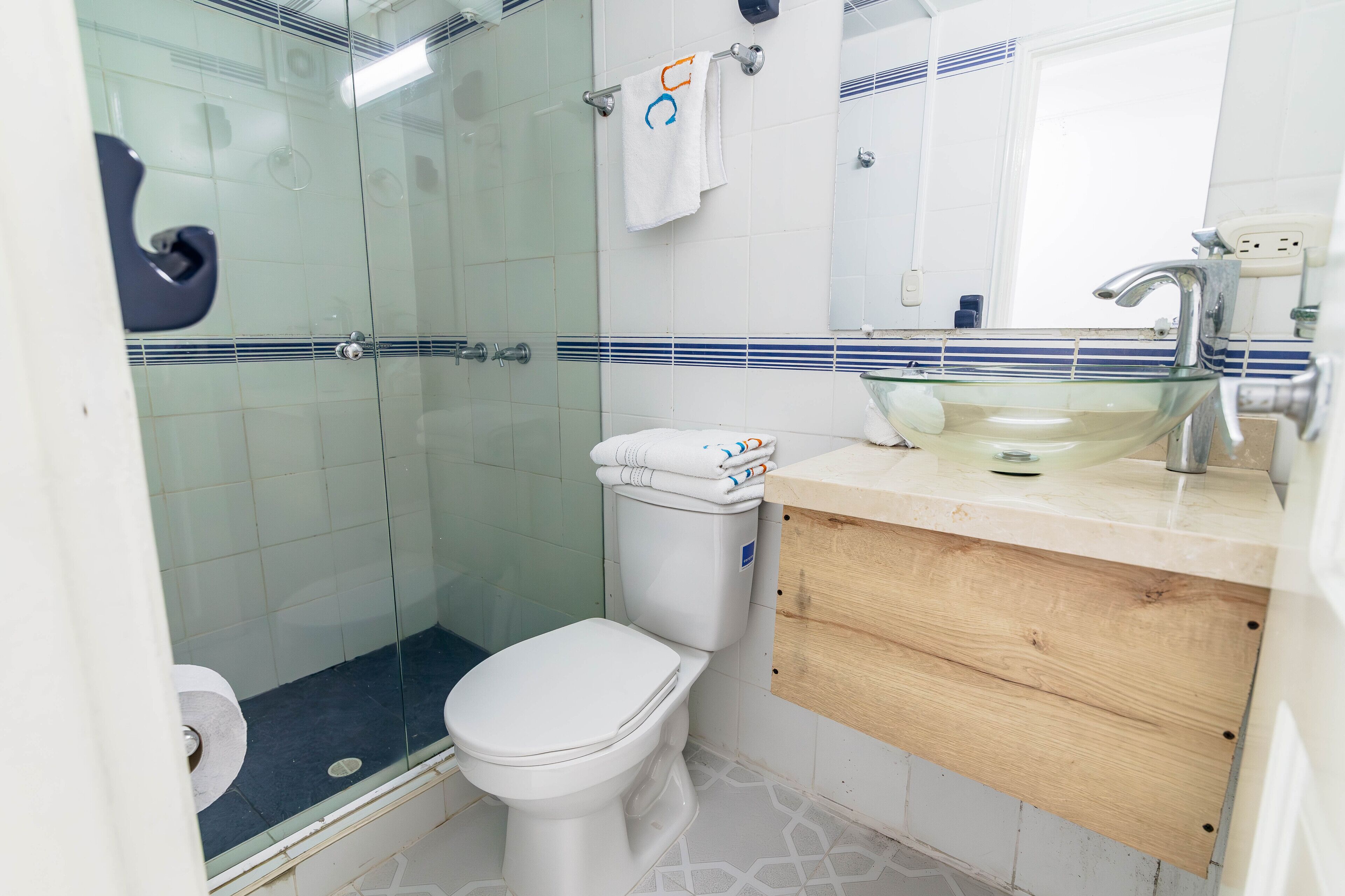Un baño con cabina de ducha de vidrio, inodoro, lavamanos con espejo y toallero con toallas.