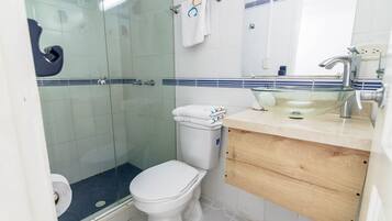 Un baño con cabina de ducha de vidrio, inodoro, lavamanos con espejo y toallero con toallas.