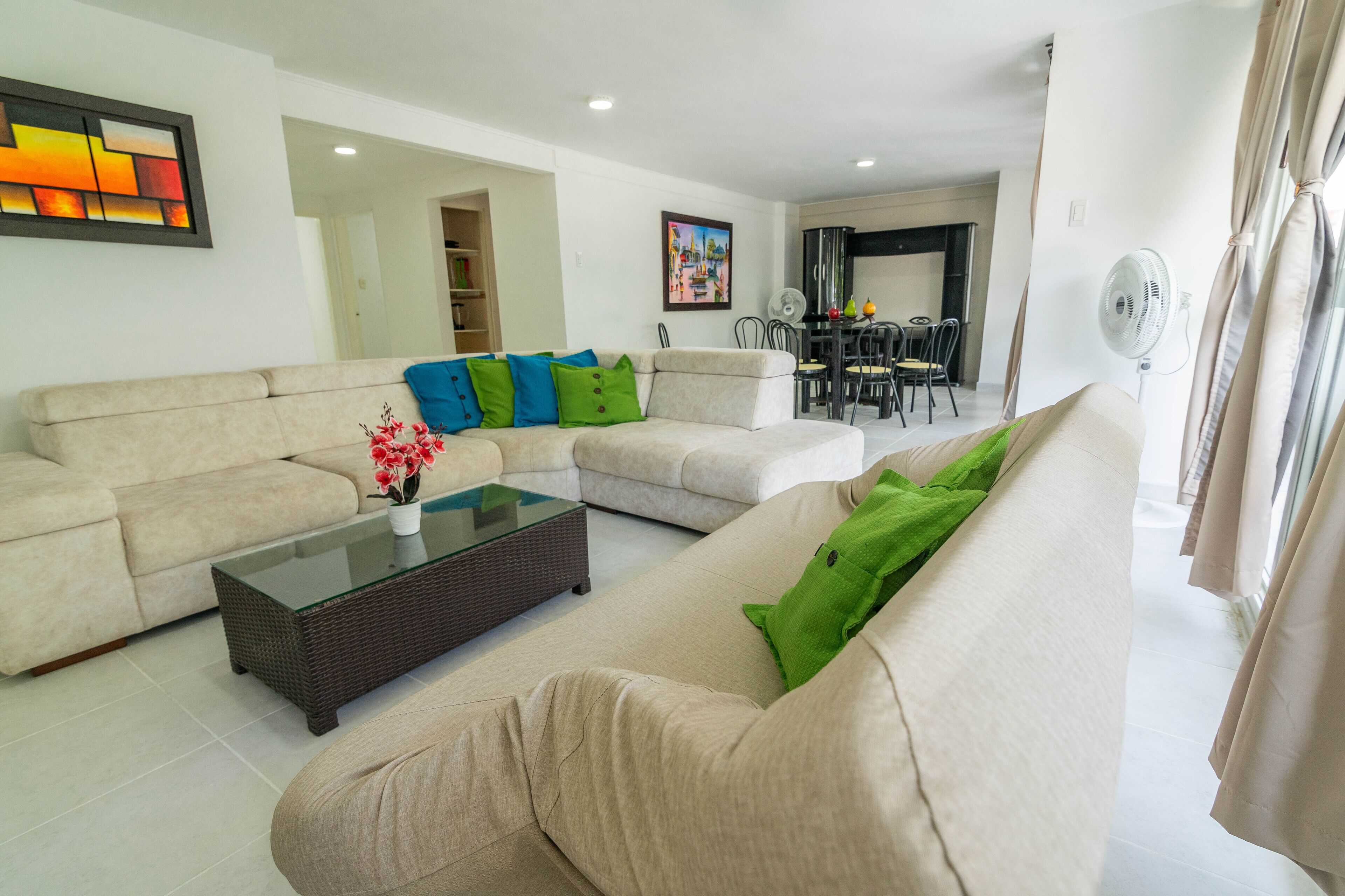 A modern living room with a beige sofa, a glass coffee table, and a dining area in the background.