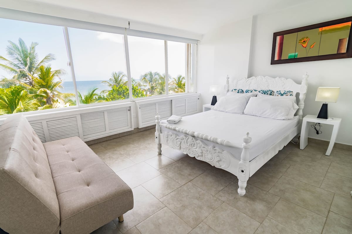 A bedroom with a bed, a nightstand, a lamp, a sofa, and a view of the ocean.